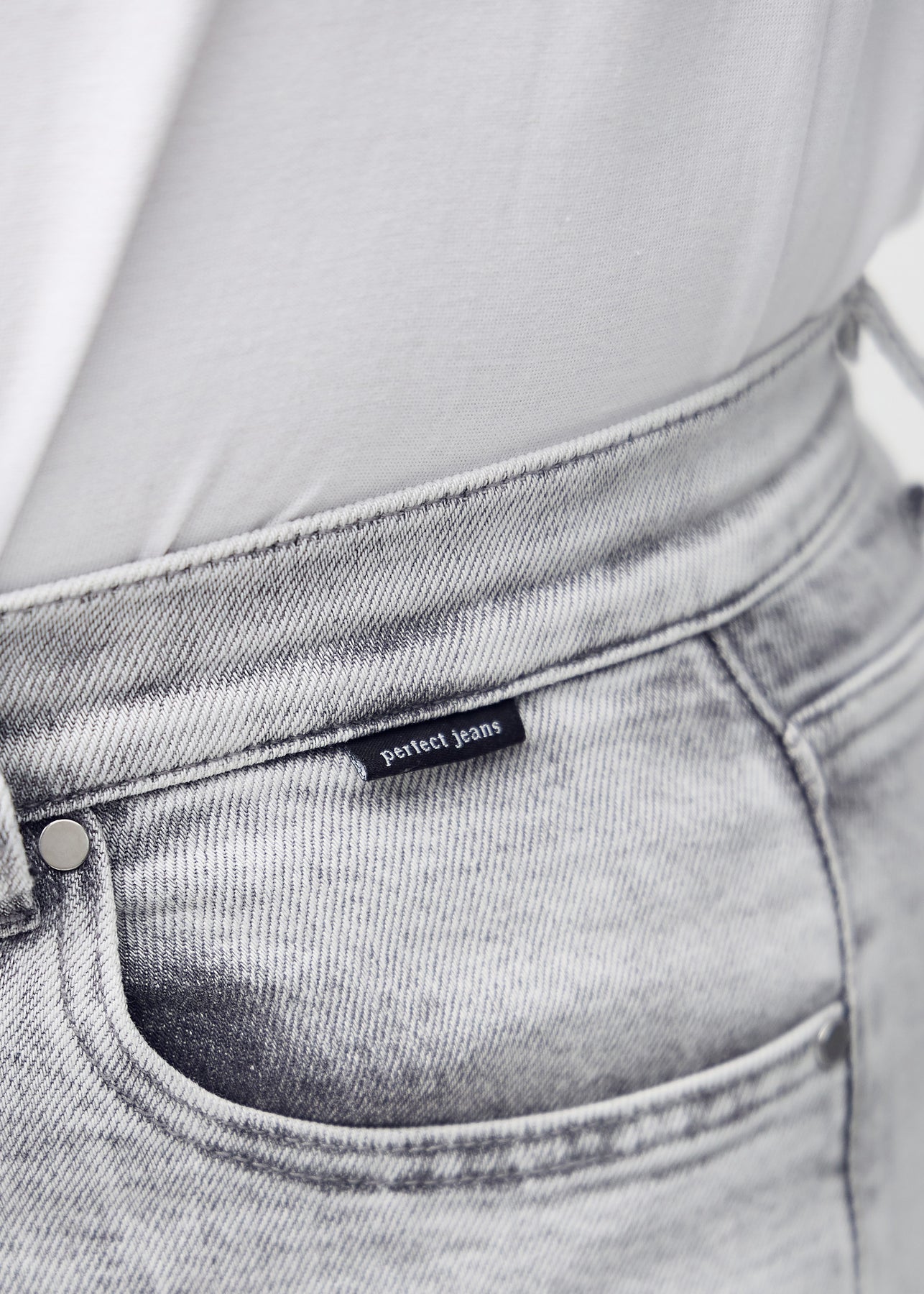 Nærbillede af lysegrå denim med Perfect Jeans logoetiket ved forlommekanten.