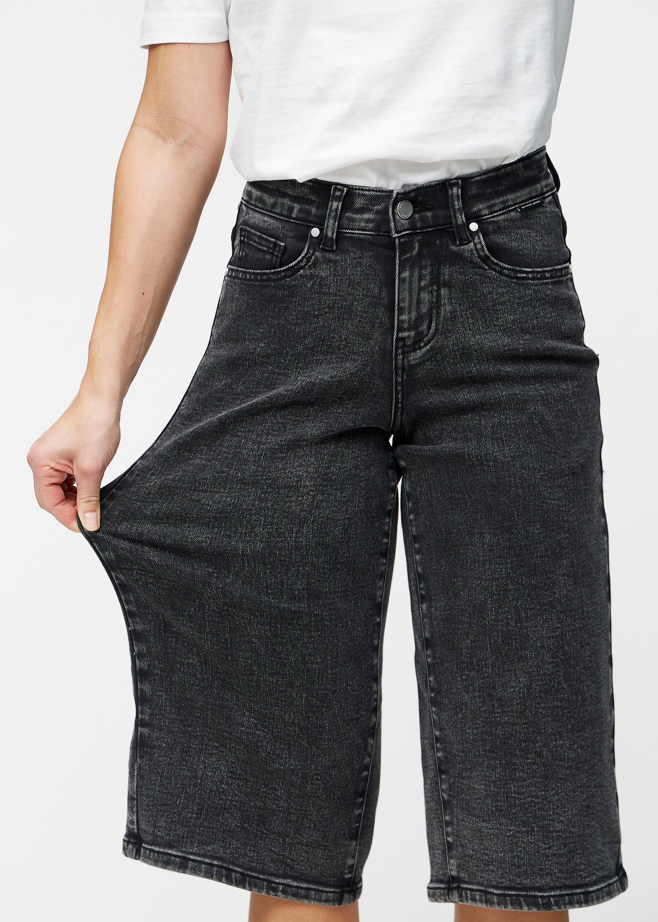 Model strækker de løse, mørkegrå denim capris i farven Shadows ved hoften for at vise materialets stretch.