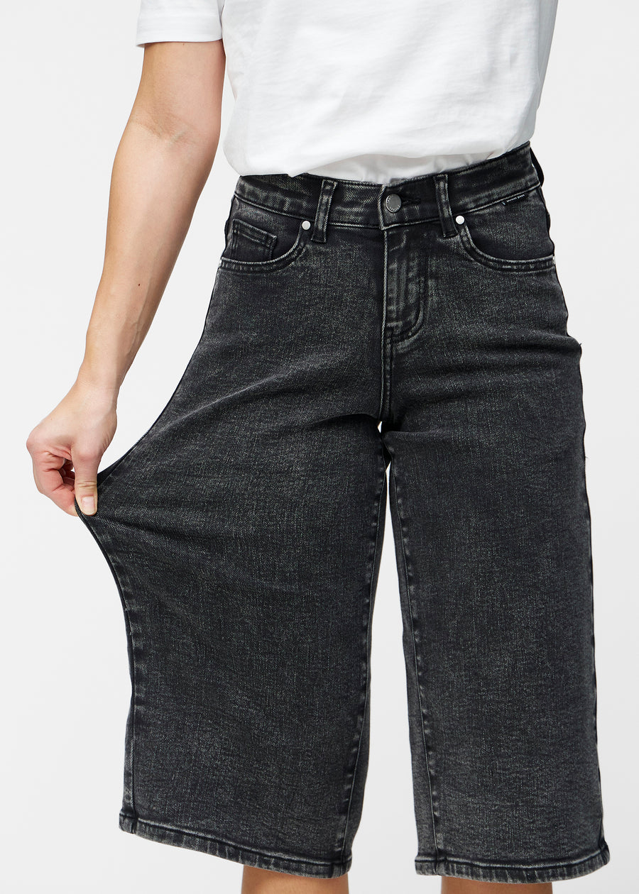 Model strækker de løse, mørkegrå denim capris i farven Shadows ved hoften for at vise materialets stretch.