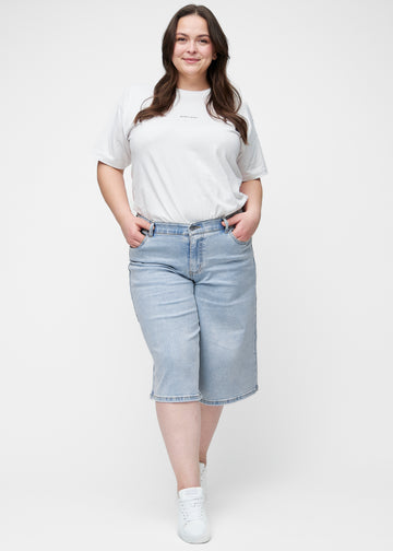 Fullt bilde av plus-size modell i løse, lyseblå denim capris i fargen Waves som går til rett under kneet.