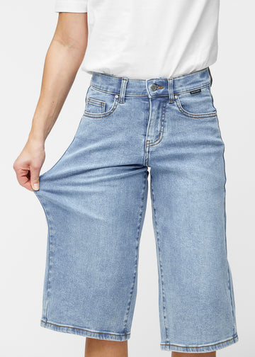 Modell strekker de løse, lyseblå denim capris i fargen Waves ved hoften for å vise materialets stretch.