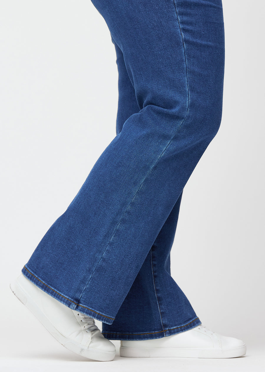 Mørkeblå mid-rise bootcut-jeans i fargen Oceans. Nærbilde ved ankelen på en plus size-modell som viser den lette utsvingen nederst.