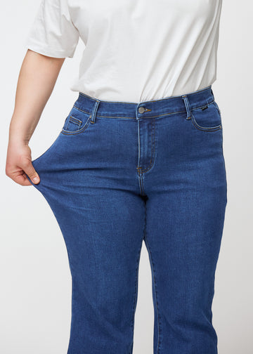 Mørkeblå mid-rise bootcut-jeans i fargen Oceans. En plus size-modell strekker jeansstoffet ved låret for å vise stretchen.