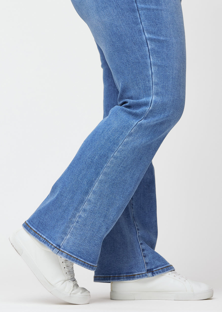 Blå mid-rise bootcut-jeans i fargen Rivers. Nærbilde ved ankelen på en plus size-modell som viser den lette utsvingen nederst.