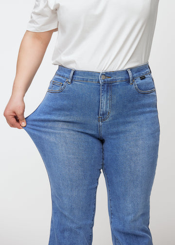Blå mid-rise bootcut-jeans i fargen Rivers. En plus size-modell strekker jeansstoffet ved låret for å vise stretchen.