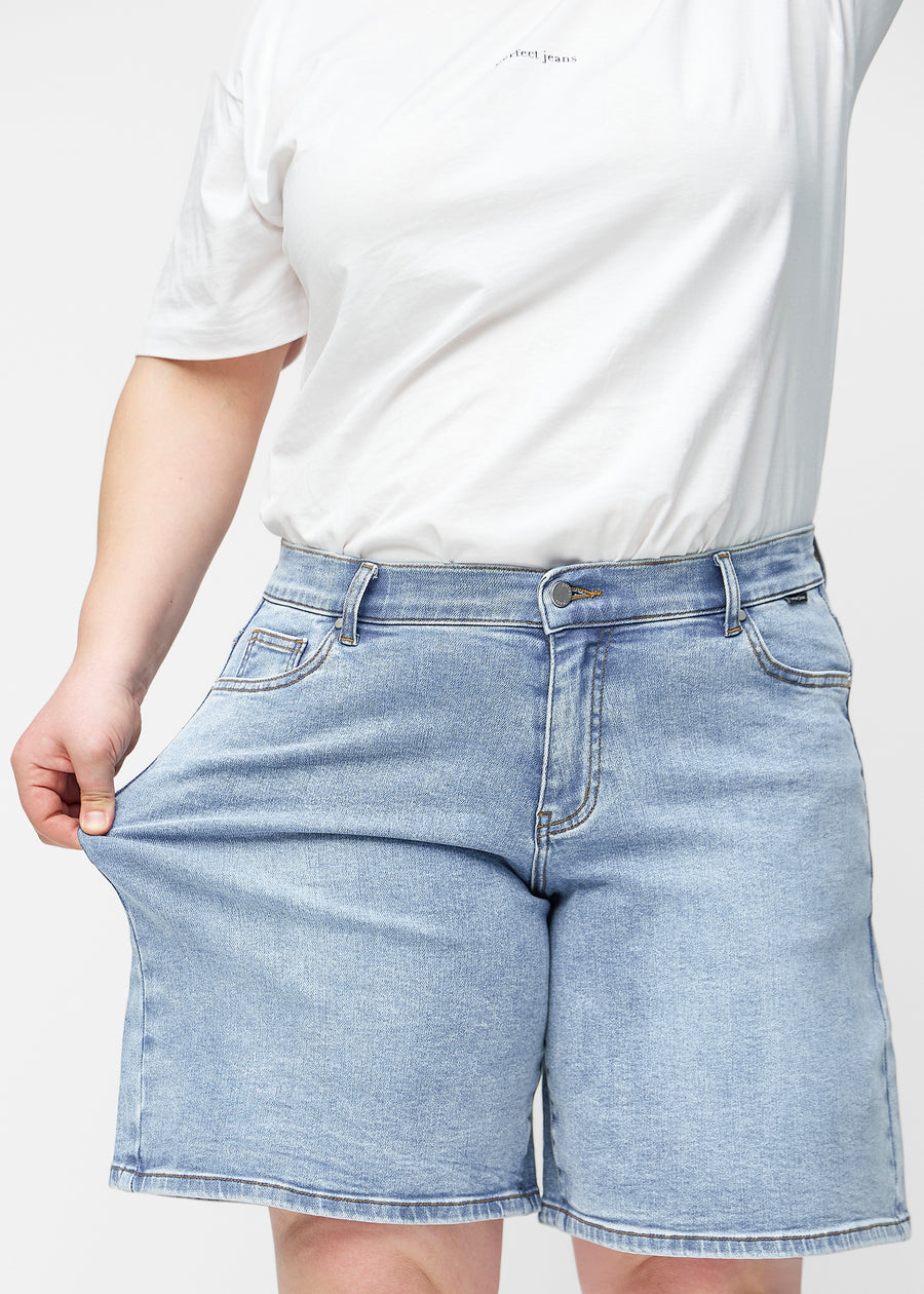 Plus-size modell strekker de løse, lyseblå denimshortsene i fargen Waves ved hoften for å vise materialets stretch.
