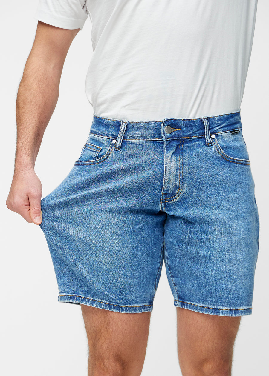 Modell strekker blå denimshorts til herre i fargen Rivers ved låret for å vise materialets stretch.