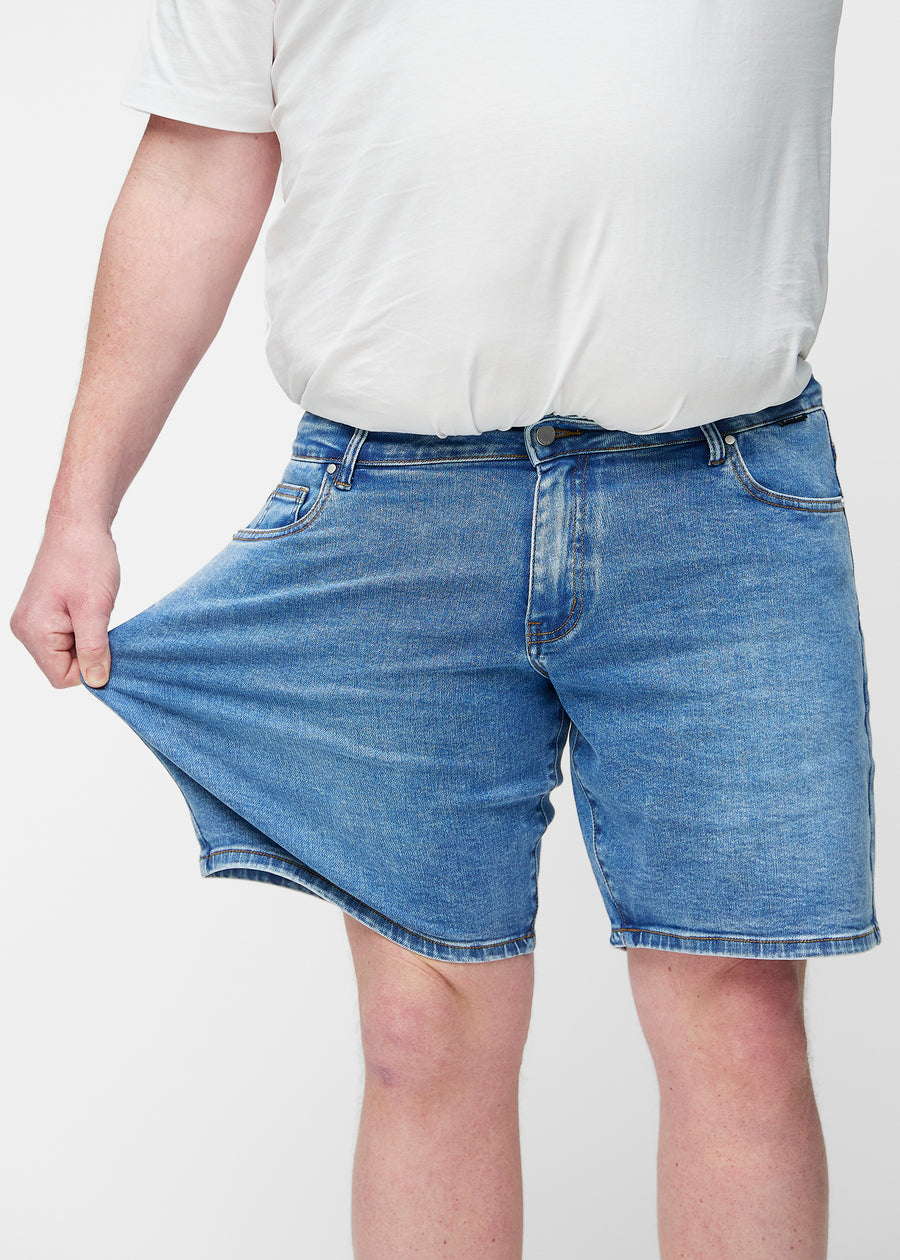 Plus-size modell strekker blå denimshorts til herre i fargen Rivers ved låret for å vise materialets stretch.
