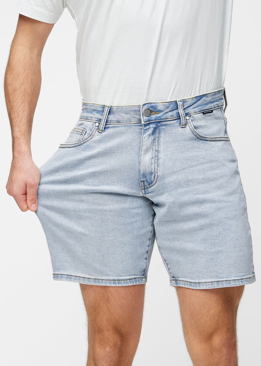 Modell strekker lyseblå denimshorts til herre i fargen Waves ved låret for å vise materialets stretch.