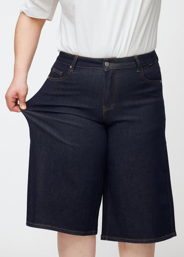 Plus-size modell strekker dyp mørkeblå denim capris til dame i fargen Midnights ved låret for å vise materialets stretch.