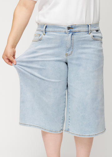 Plus-size-modell strekker de løse, lyseblå Waves denimcapribuksene ved hoften for å vise materialets stretch.