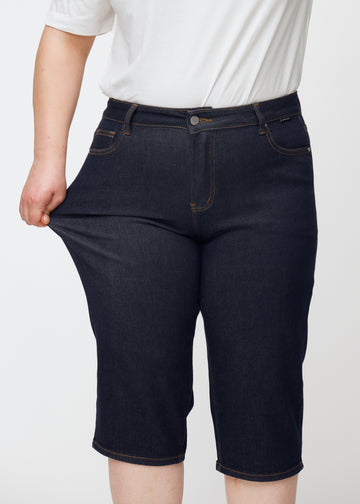 Plus-size modell strekker dyp mørkeblå denim capris til dame i fargen Midnights ved låret for å vise stretch.