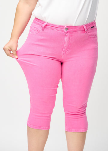 Skinny-capris til kvinner i pastellrosa jeansstoff, fargen Flamingos. En plus size-modell strekker stoffet for å vise fleksibiliteten.