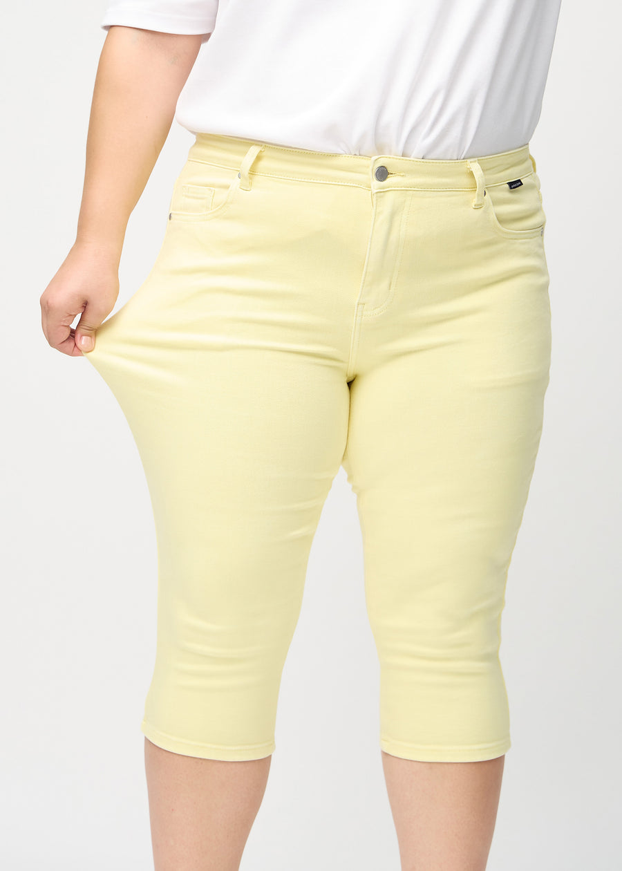 Skinny-capris til kvinner i pastellgult jeansstoff, fargen Lemonades. En plus size-modell strekker stoffet for å vise fleksibiliteten.