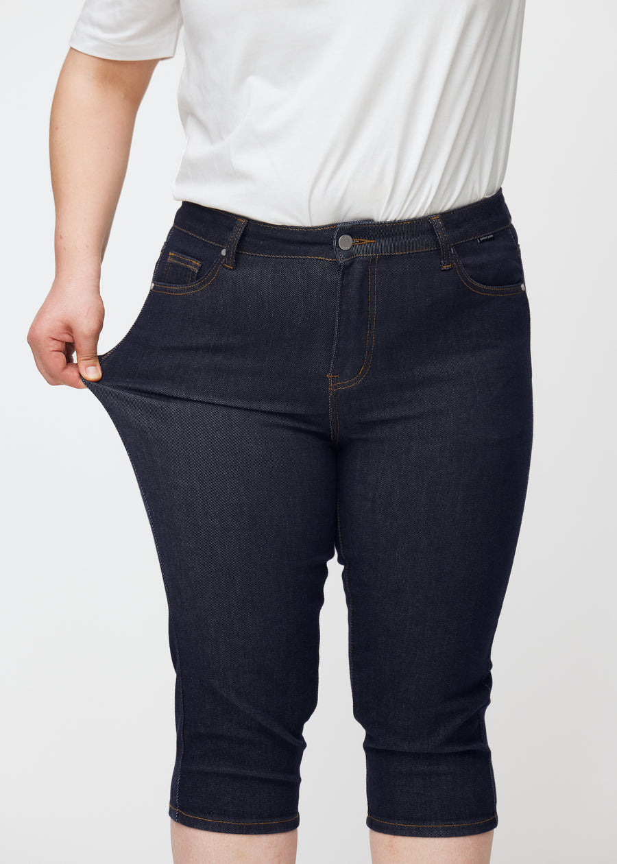Plus-size modell strekker dyp mørkeblå denim capris til dame i fargen Midnights ved låret for å vise stretch.