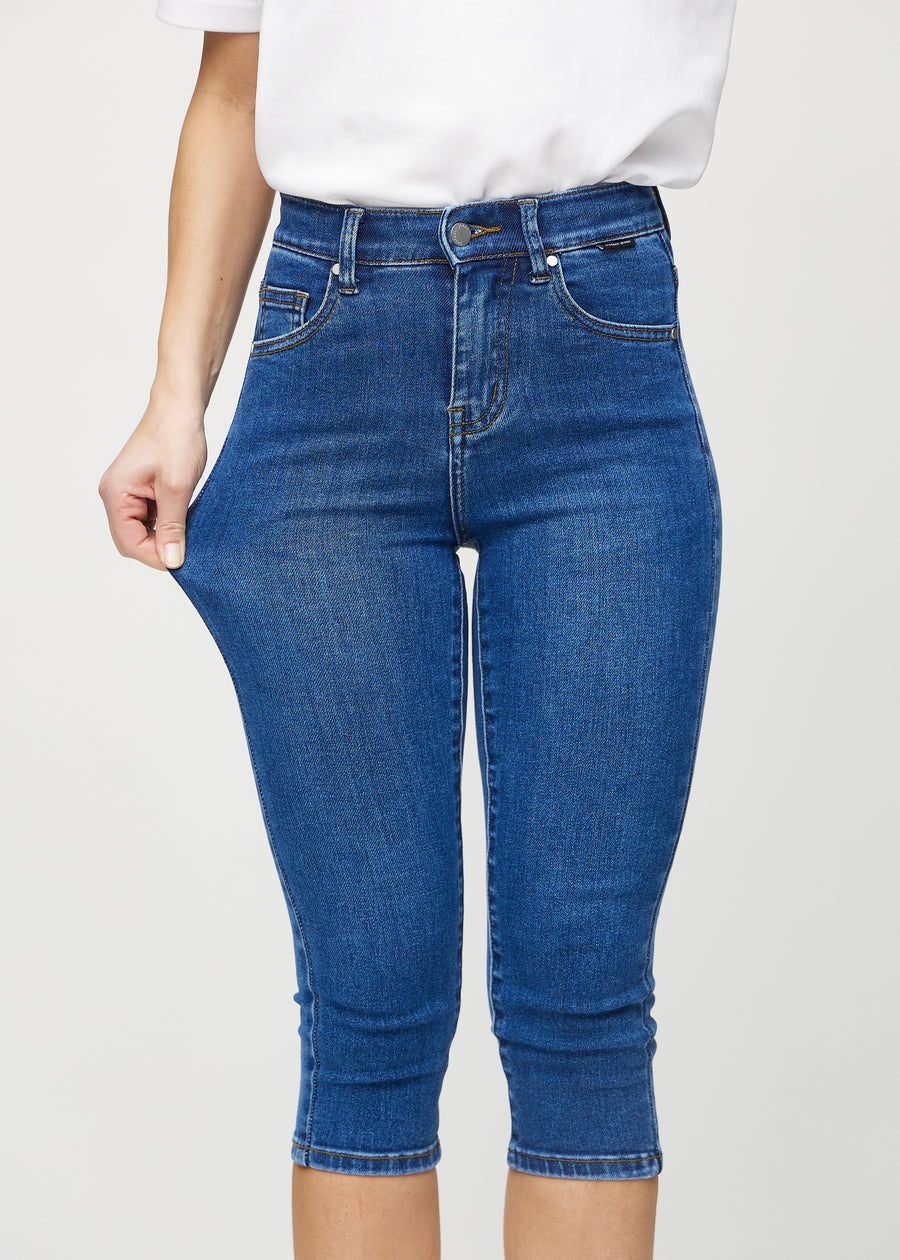 Skinny-capris til kvinner i mørkeblå denim, fargen Oceans. En modell strekker stoffet for å vise fleksibiliteten.