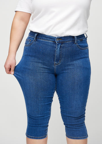 Skinny-capris til kvinner i mørkeblå denim, fargen Oceans. En plus size-modell strekker stoffet for å vise fleksibiliteten.