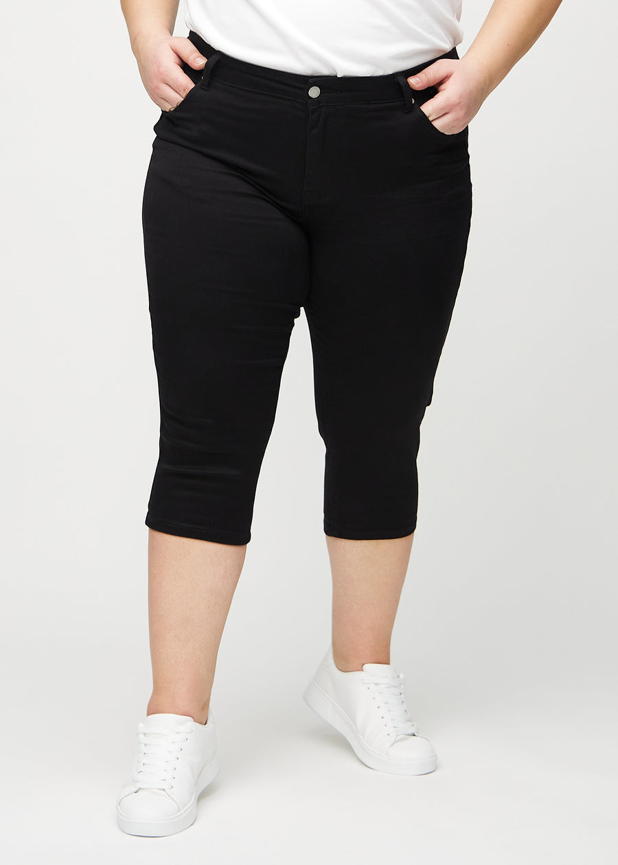 Svarte skinny capris, modellnavn Ravens, som sitter tett til benet og slutter rett under kneet på en plus-size-modell, sett forfra.