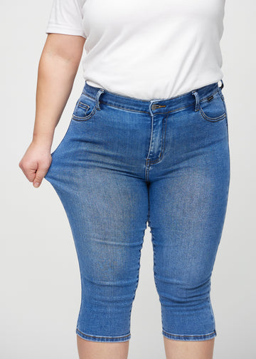 Skinny-capris til kvinner i blå denim, fargen Rivers. En plus size-modell strekker stoffet for å vise fleksibiliteten.