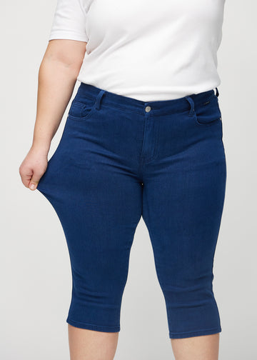 Skinny-capris til kvinner i mørkeblått jeansstoff, fargen Royals. En plus size-modell strekker stoffet for å vise fleksibiliteten.