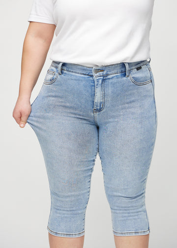 Skinny-capris til kvinner i lyseblå denim, fargen Waves. En plus size-modell strekker stoffet for å vise fleksibiliteten.