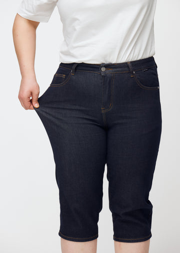 Plus-size modell strekker dyp mørkeblå denim capris til dame i fargen Midnights ved låret for å vise stretch.
