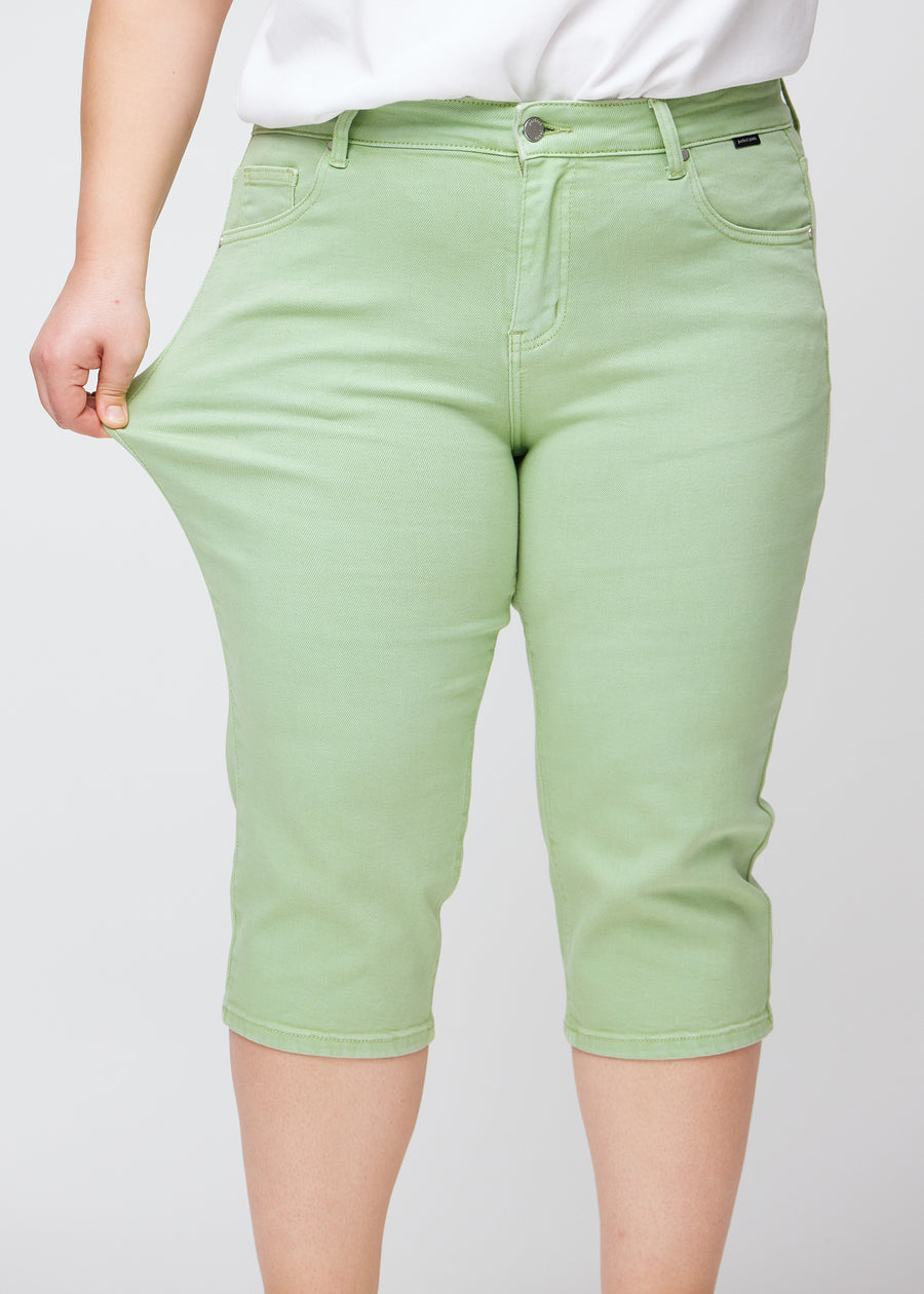 Slim-capris til kvinner i pastelllysegrønt jeansstoff, fargen Mints. En plus size-modell strekker stoffet ved låret for å vise stretch og fleksibilitet.