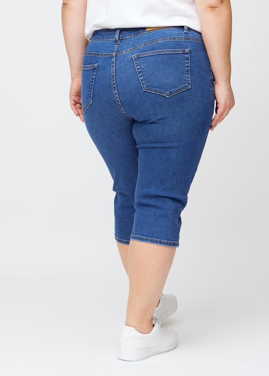 Slim-capris til kvinner i mørkeblå denim, fargen Oceans. Sett bakfra på en plus size-modell, med fokus på passform, lengde og baklommer.