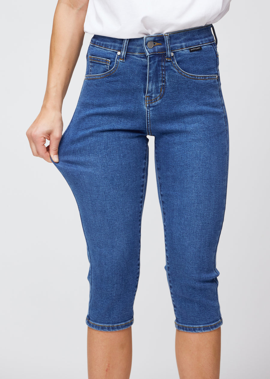 Slim-capris til kvinner i mørkeblå denim, fargen Oceans. En modell strekker stoffet ved låret for å vise stretch og fleksibilitet.