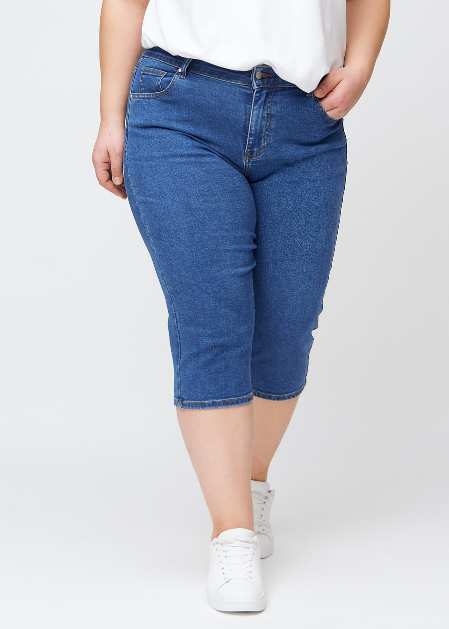 Slim-capris til kvinner i mørkeblå denim, fargen Oceans. Sett forfra på en plus size-modell, med figursydd passform til rett under kneet.