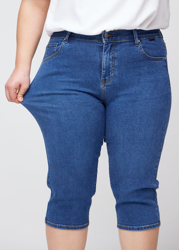Slim-capris til kvinner i mørkeblå denim, fargen Oceans. En plus size-modell strekker stoffet ved låret for å vise stretch og fleksibilitet.