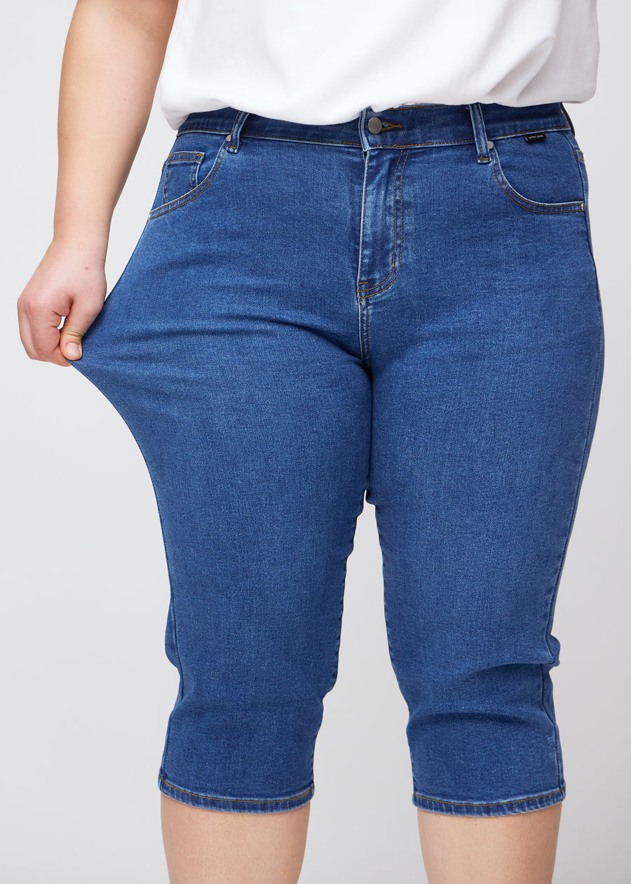 Slim-capris til kvinner i mørkeblå denim, fargen Oceans. En plus size-modell strekker stoffet ved låret for å vise stretch og fleksibilitet.
