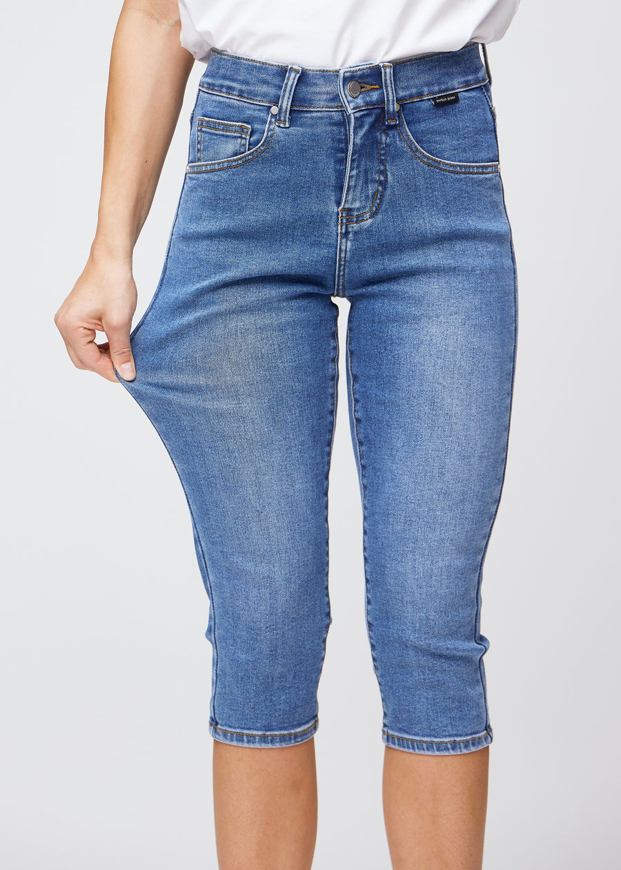 Slim-capris til kvinner i blå denim, fargen Rivers. En modell strekker stoffet ved låret for å vise stretch og fleksibilitet.