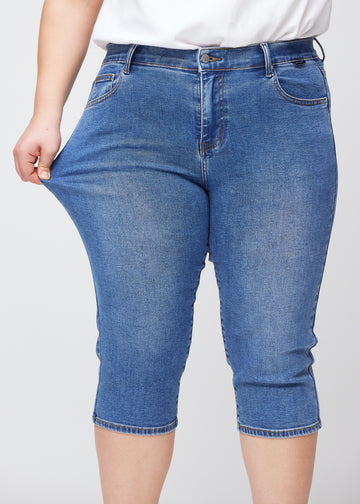 Slim-capris til kvinner i blå denim, fargen Rivers. En plus size-modell strekker stoffet ved låret for å vise stretch og fleksibilitet.