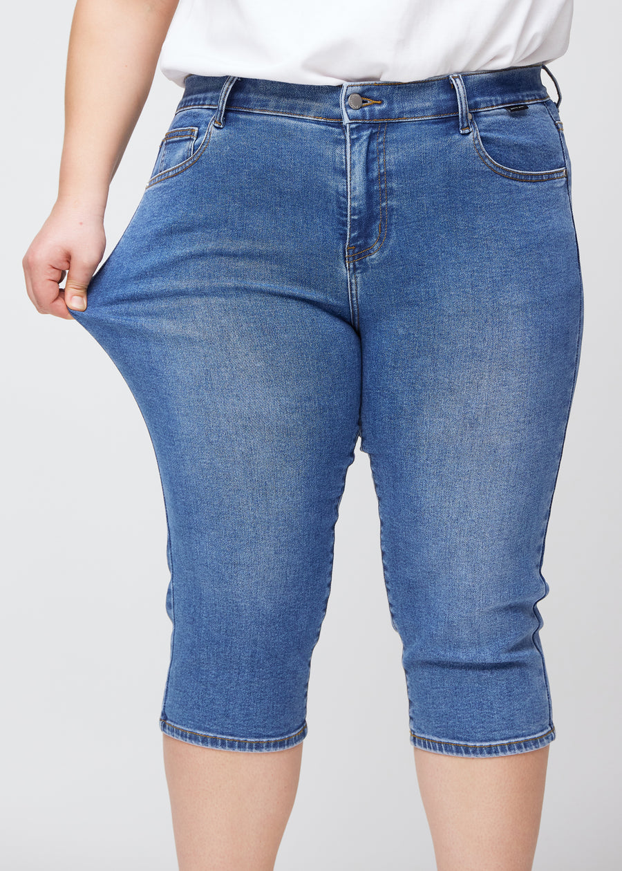 Slim-capris til kvinner i blå denim, fargen Rivers. En plus size-modell strekker stoffet ved låret for å vise stretch og fleksibilitet.