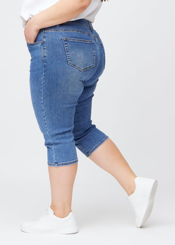 Slim-capris til kvinner i blå denim, fargen Rivers. Sett fra siden på en plus size-modell, med figursydd passform og mellomhøyt liv (Mid Rise).
