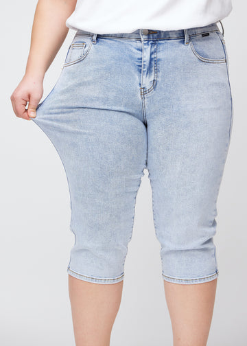Slim-capris til kvinner i lyseblå denim, fargen Waves. En plus size-modell strekker stoffet ved låret for å vise stretch og fleksibilitet.