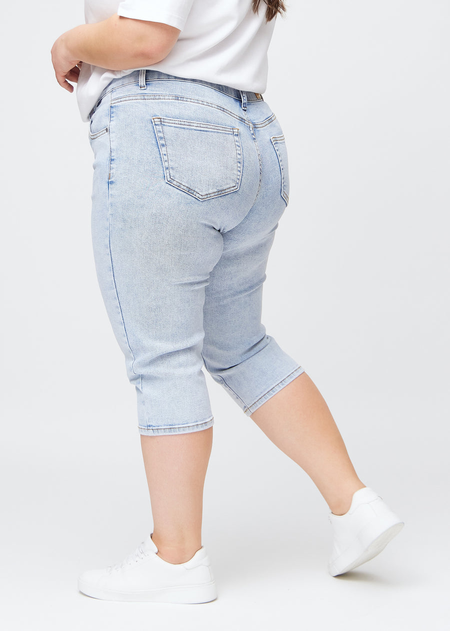 Slim-capris til kvinner i lyseblå denim, fargen Waves. Sett fra siden på en plus size-modell, med figursydd passform og mellomhøyt liv (Mid Rise).
