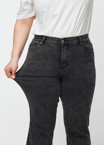 Mørkegrå mid-rise bootcut-jeans i fargen Shadows. En plus size-modell strekker jeansstoffet ved låret for å vise stretchen.