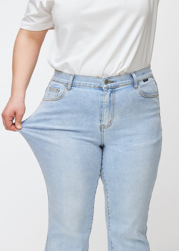 Lyseblå mid-rise bootcut-jeans i fargen Waves. En plus size-modell strekker jeansstoffet ved låret for å vise stretchen.