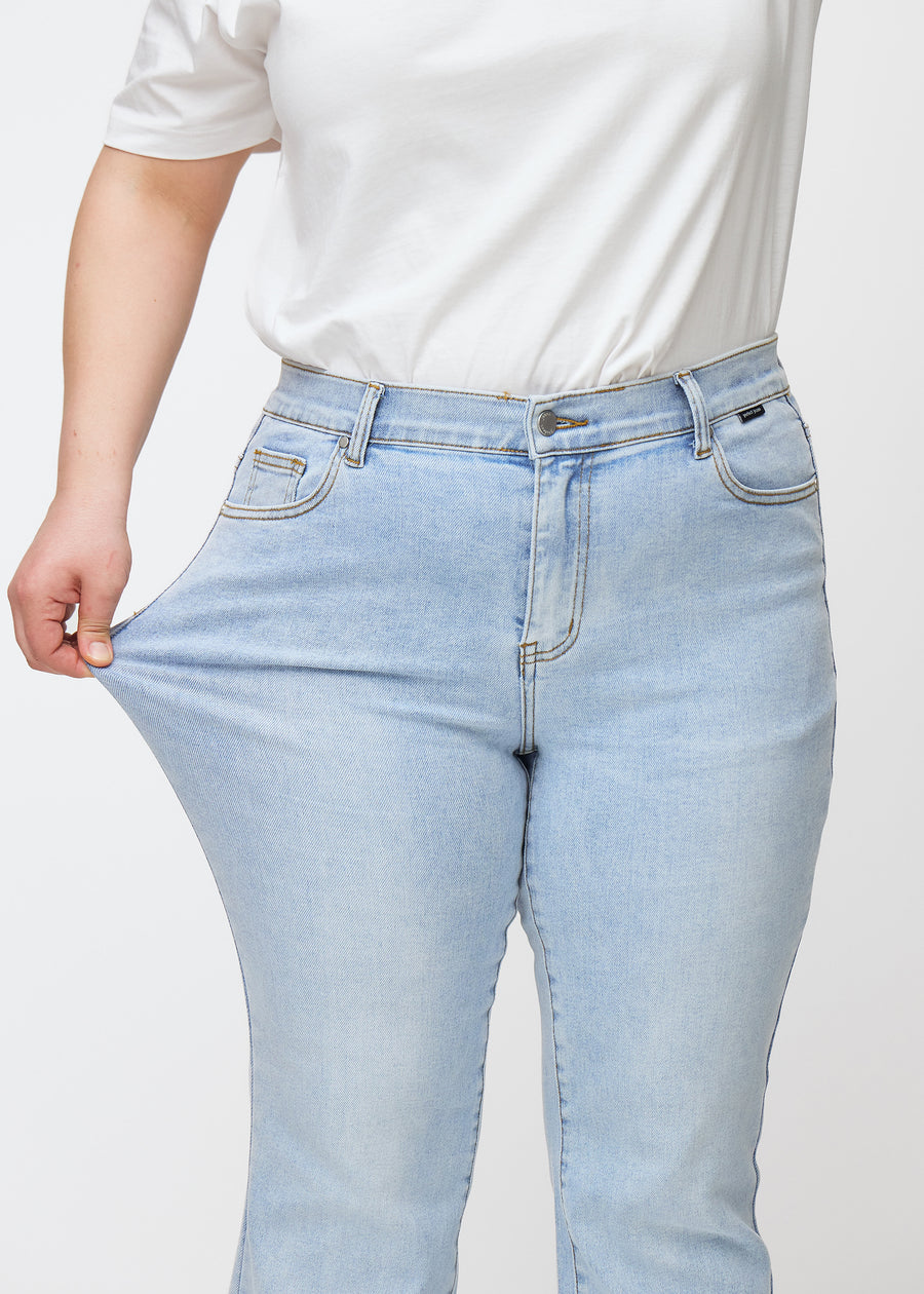 Lyseblå mid-rise bootcut-jeans i fargen Waves. En plus size-modell strekker jeansstoffet ved låret for å vise stretchen.