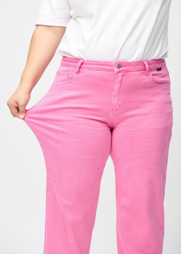 Loose-jeans til kvinner i lyserosa, fargen Flamingos. En plus size-modell strekker jeansstoffet ved låret for å vise stretchen.