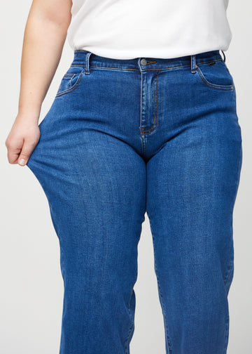 Loose-jeans til kvinner i mørkeblå, fargen Oceans. En plus size-modell strekker jeansstoffet ved låret for å vise stretchen.