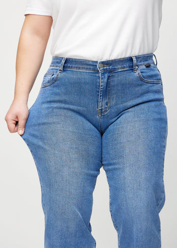 Loose-jeans til kvinner i blå, fargen Rivers. En plus size-modell strekker jeansstoffet ved låret for å vise stretchen.