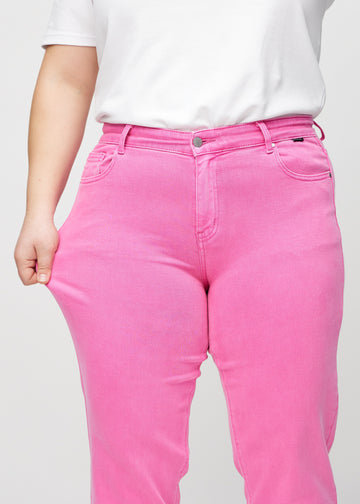 Regular-jeans til kvinner i lyserosa, fargen Flamingos. En plus size-modell strekker stoffet ved låret for å vise stretchen.