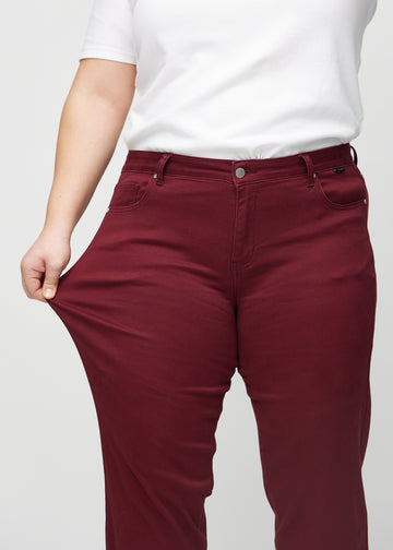 Regular-jeans til kvinner i mørkerød, fargen Santas. En plus size-modell strekker stoffet ved låret for å vise stretchen.