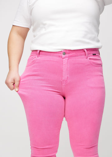Skinny-jeans til kvinner i lyserosa, fargen Flamingos. En plus size-modell strekker jeansstoffet ved låret for å vise stretchen.