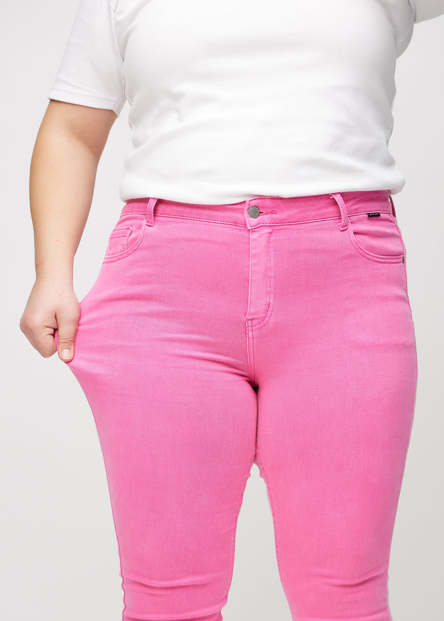 Skinny-jeans til kvinner i lyserosa, fargen Flamingos. En plus size-modell strekker jeansstoffet ved låret for å vise stretchen.