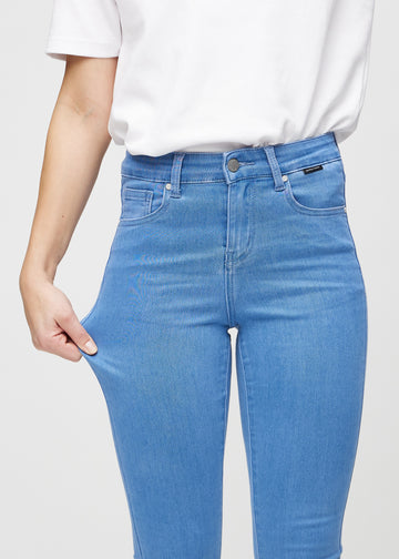 Skinny-jeans til kvinner i blå, fargen Geraniums. En modell strekker jeansstoffet ved låret for å vise stretchen.