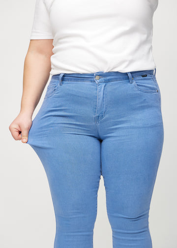 Skinny-jeans til kvinner i blå, fargen Geraniums. En plus size-modell strekker jeansstoffet ved låret for å vise stretchen.